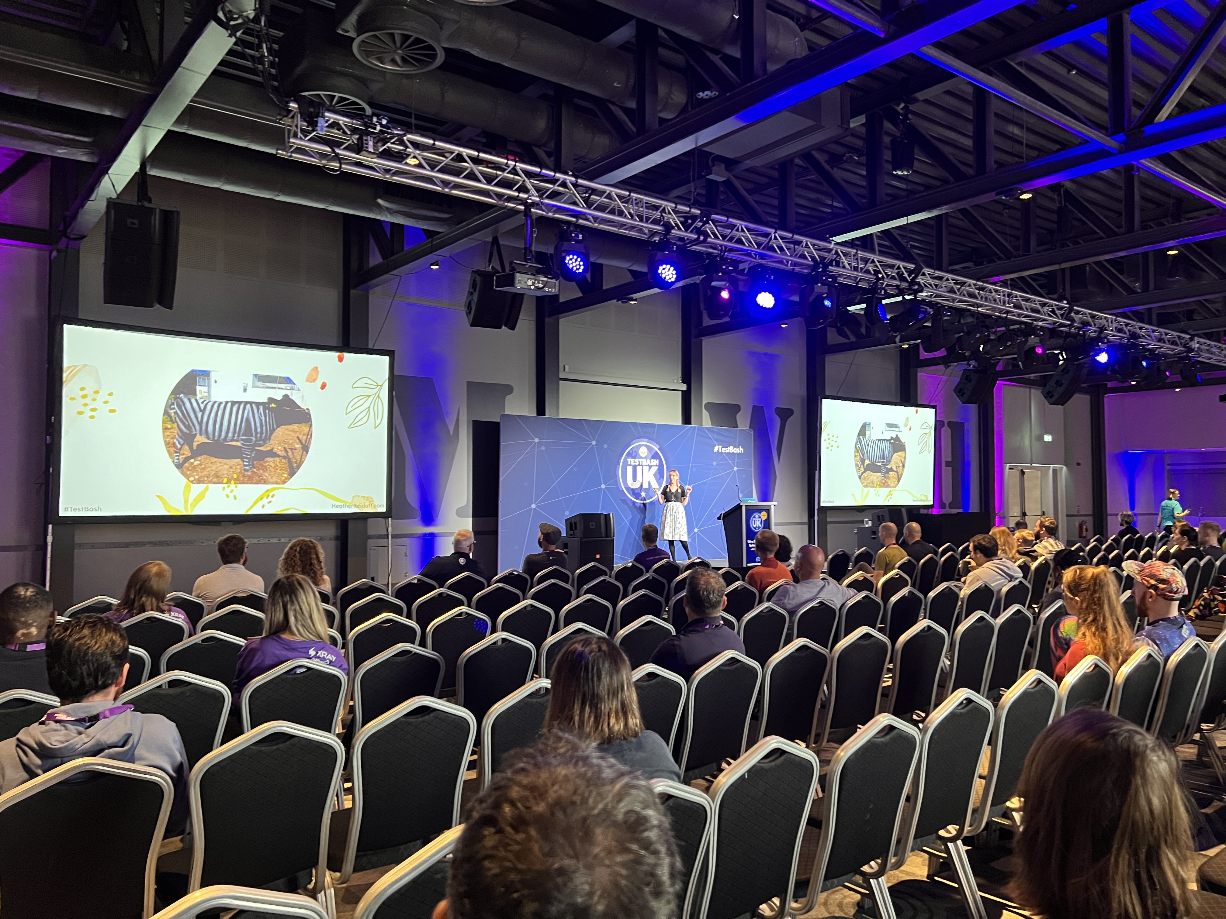 A conference room full of many people. There is a woman on stage presenting a talk. 