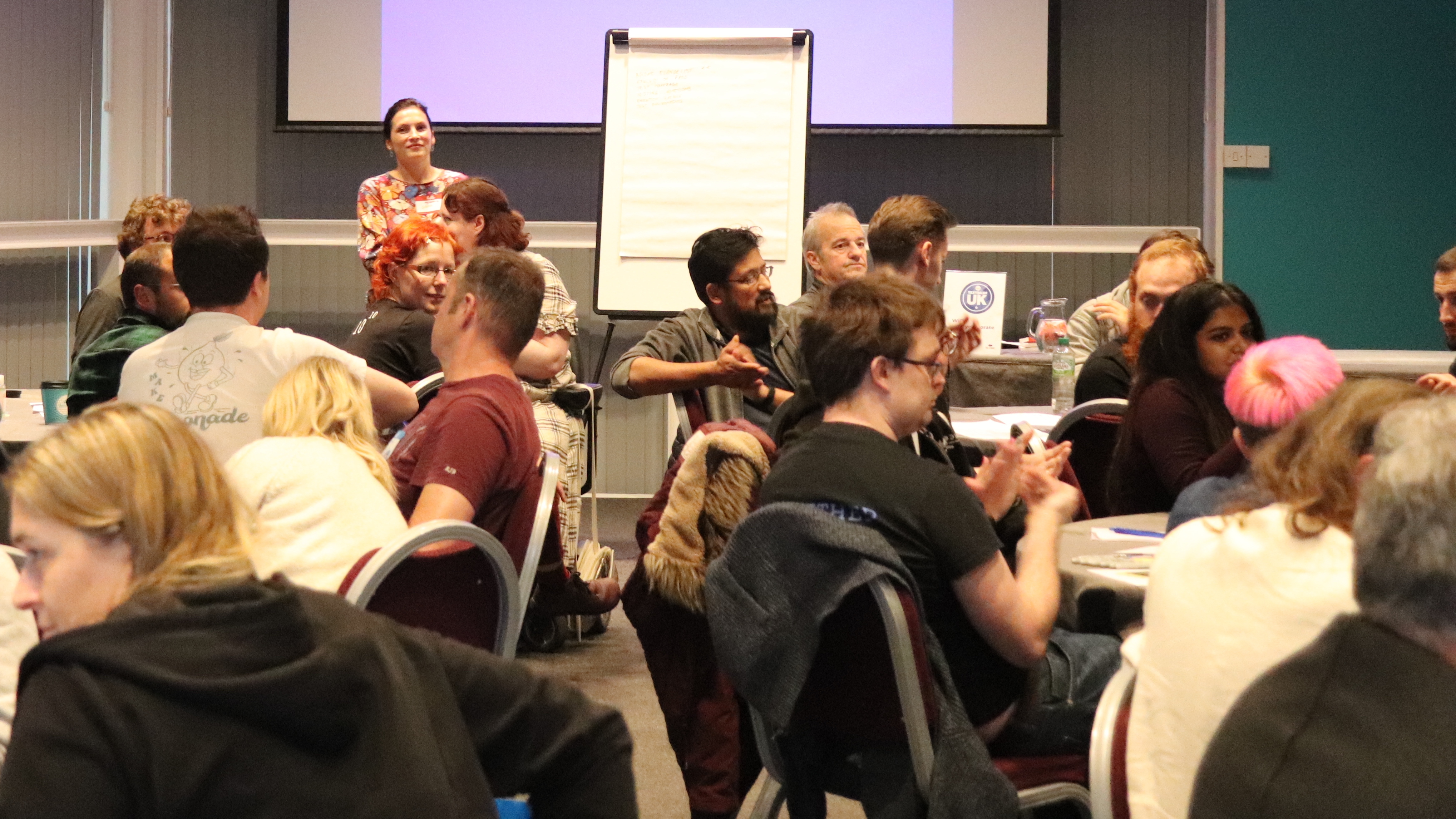 A group of people sat at desks in a conference. There is a woman stood up running a workshop.
