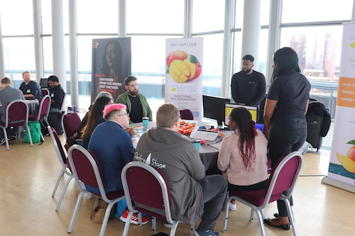 A group of people sat at desks in a conference. There are two people stood up running an activity.