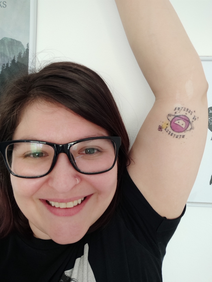 A photo of a smiling woman with her arm raised. The arm has a Ministry of Testing logo tattoo on it.